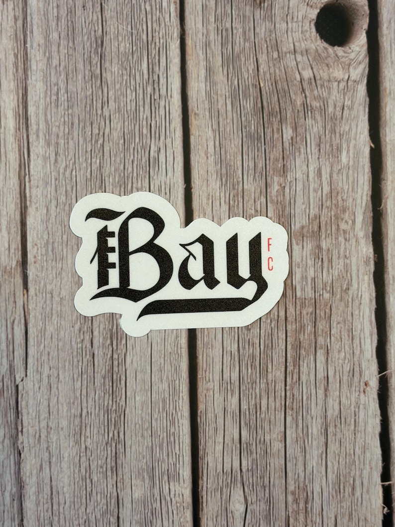 Puede incluir: Una pegatina blanca con la palabra "Bay" en fuente g&oacute;tica negra, con "FC" en rojo. La pegatina est&aacute; sobre una superficie de madera envejecida.