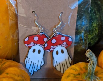 May include: A pair of red and white mushroom earrings with a ghost face on each. The earrings are on a brown card with a hole in the center.