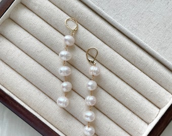 Può includere: Un paio di orecchini d'oro con gocce di perle bianche. Gli orecchini sono esposti in una scatola per gioielli foderata in tessuto bianco con cornice in legno marrone.