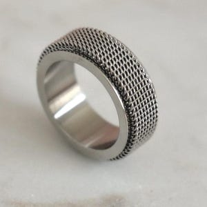 May include: A silver-colored ring with a woven mesh design around the band. The ring has a smooth, polished interior and a textured exterior. The ring is a simple, yet stylish piece of jewelry.
