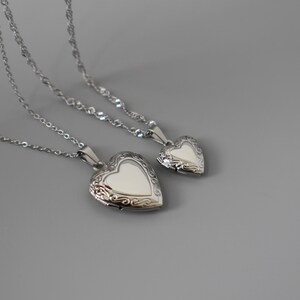 Two silver heart-shaped locket pendants with intricate designs are displayed on a gray background.