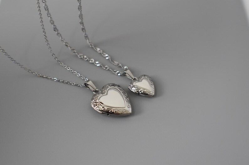 Two silver heart-shaped locket pendants with intricate designs are displayed on a gray background.