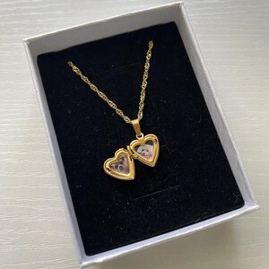 A gold-colored locket pendant with a heart-shaped compartment, displayed in a white jewelry box.