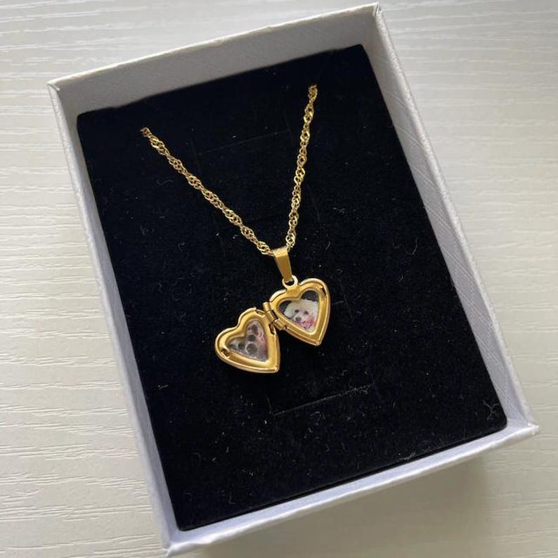 A gold-colored locket pendant with a heart-shaped compartment, displayed in a white jewelry box.