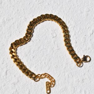 A gold-colored bracelet with a clasp and a small tag attached to it, lying on a white textured surface.