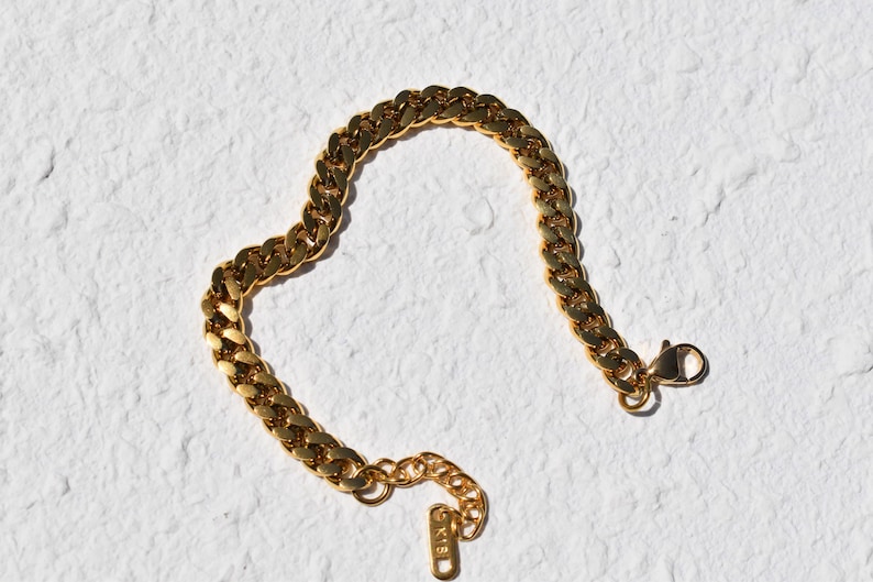 A gold-colored bracelet with a clasp and a small tag attached to it, lying on a white textured surface.