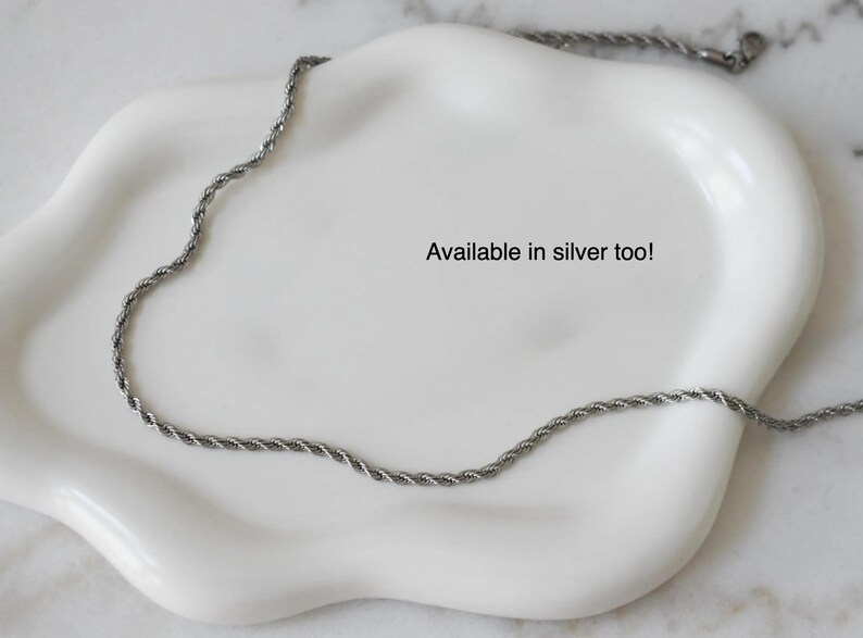 A silver necklace chain is displayed on a white plate, with the text "Available in silver too!" written on the plate.