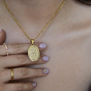 May include: Gold-toned necklace with an oval pendant featuring the Pisces symbol. The pendant hangs from a delicate chain. The image also shows gold rings on a hand with manicured nails.