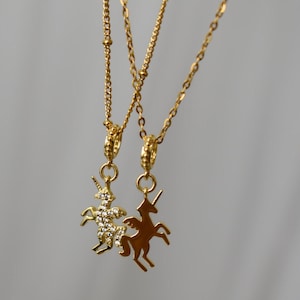 May include: Two gold necklaces with unicorn pendants. One pendant is a silhouette, the other is encrusted with small, clear crystals. The chains are delicate, hanging against a neutral background.