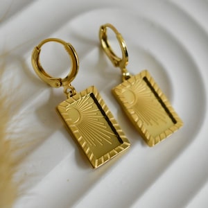 May include: A pair of gold-colored hoop earrings. Each earring features a rectangular pendant with a sunburst design and a crescent moon. The earrings are displayed on a white surface.