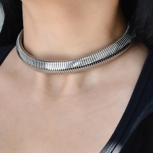 May include: A silver-colored choker necklace. The necklace is made of a series of connected, ribbed metal segments. The choker sits close to the neck and reflects light. The model is wearing a black top.