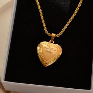 May include: A gold-colored heart-shaped locket necklace with the words "Proud of You Always" engraved on the front. The locket hangs from a gold rope chain, presented in a black box.