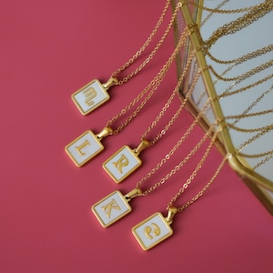 May include: Gold chain necklaces with rectangular pendants. Each pendant features a white background, a gold border, and a gold letter. The necklaces are presented on a pink surface.