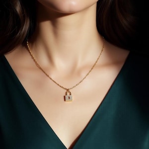 Peut inclure: Un collier doré avec un petit pendentif en forme de cadenas. Le pendentif est orné de petits cristaux transparents. Le collier est porté sur un haut vert foncé, créant un contraste qui met en valeur le bijou.