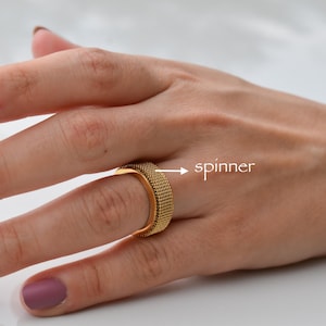 May include: A gold-colored spinner ring with a textured surface is shown on a hand. The ring is a square shape. The nails are painted with marble and glitter designs.