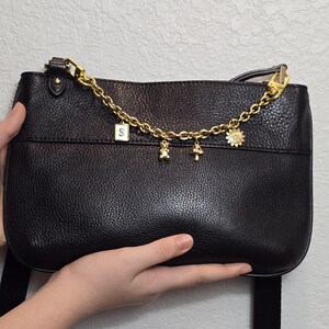 A hand holding a black leather handbag with a gold chain and charms attached to it.