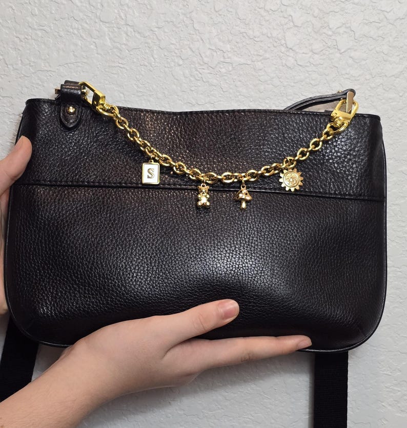 A hand holding a black leather handbag with a gold chain and charms attached to it.
