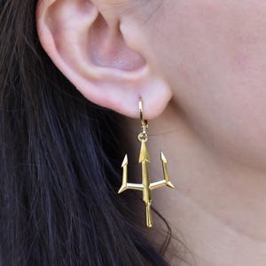 A close-up view of a person's ear wearing a pair of gold-colored earrings shaped like a trident.