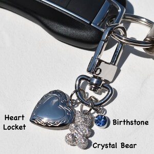 May include: Silver heart-shaped locket, crystal bear charm, and blue birthstone pendant attached to a keychain. The keychain is made of silver metal and is attached to a car key.