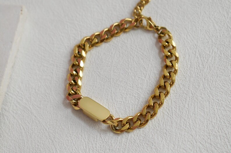 A gold-colored bracelet with a rectangular charm attached to it, resting on a white surface.