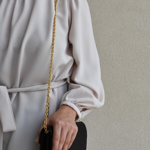 A person wearing a white blouse and holding a black purse is standing against a plain background.
