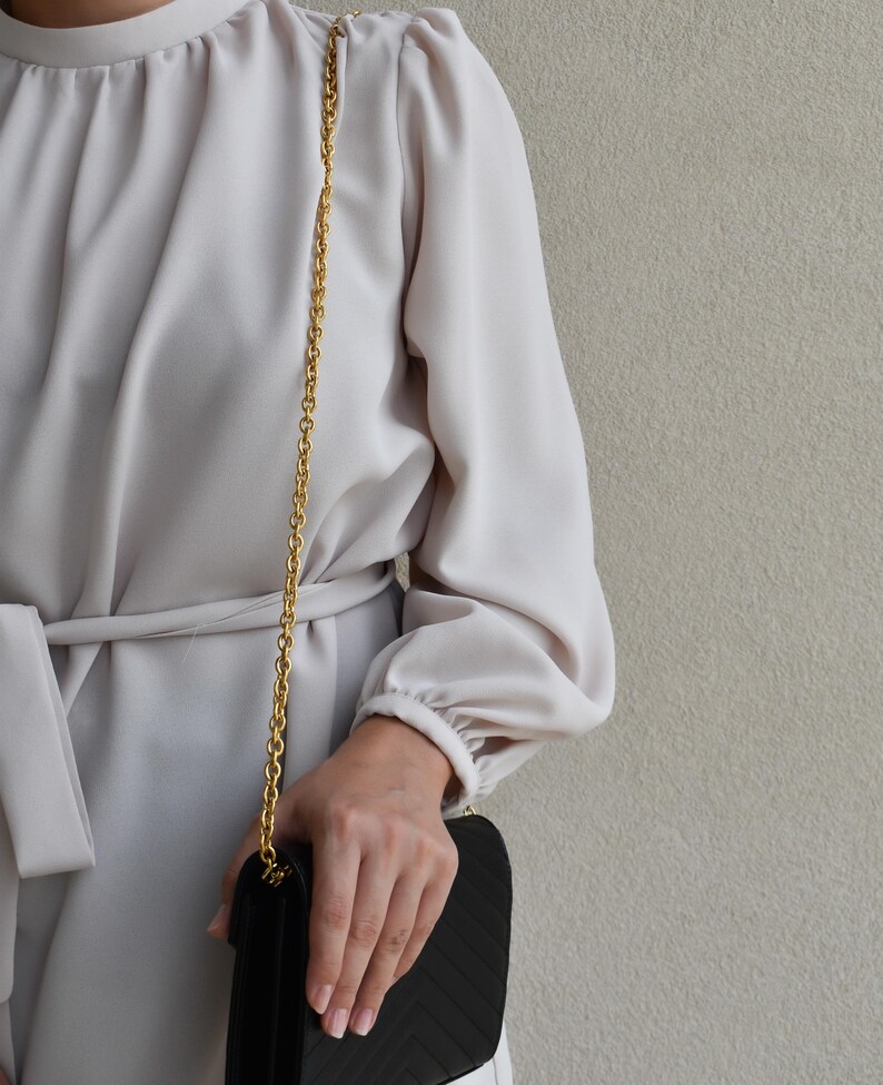 A person wearing a white blouse and holding a black purse is standing against a plain background.