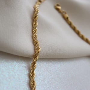 A close-up view of a gold chain necklace with a twisted design, resting on a white fabric surface.