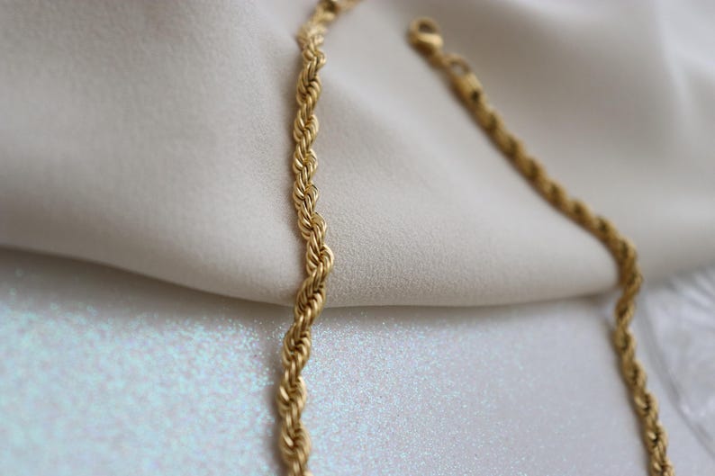 A close-up view of a gold chain necklace with a twisted design, resting on a white fabric surface.