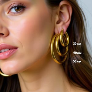 May include: Gold hoop earrings in three sizes: 30mm, 40mm, and 50mm. The earrings are displayed on a person's ear, showcasing their polished, reflective surface. The image highlights the different sizes available.