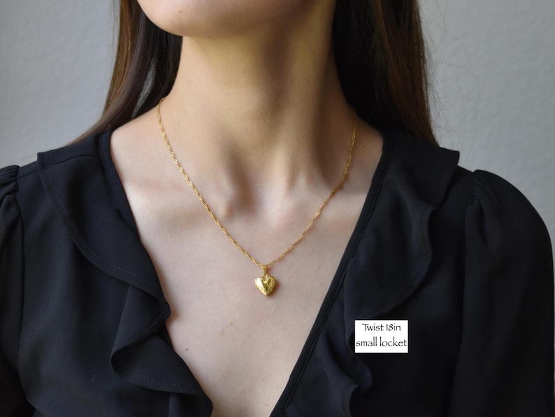 A woman wearing a black blouse with a gold necklace and a small heart-shaped pendant.
