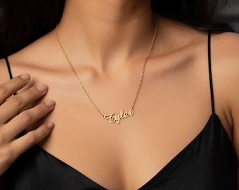 May include: A gold-colored necklace with the name "Taylor" in cursive script. The necklace is worn against a black top. The chain is delicate, and the name is the focal point of the jewelry.