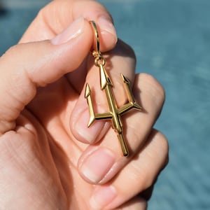 A gold-colored trident pendant is being held in a person's hand, with the background blurred to focus attention on the pendant.