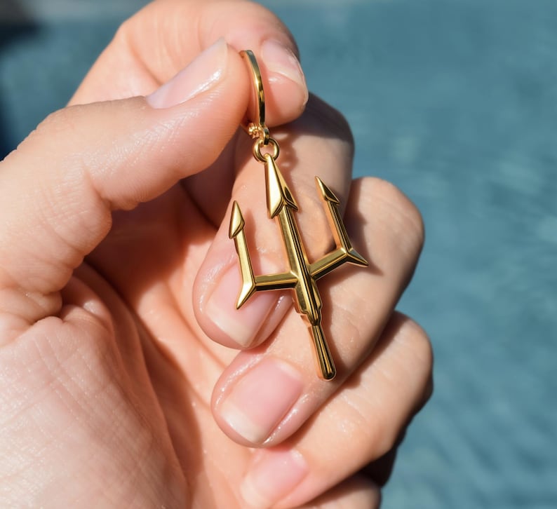 A gold-colored trident pendant is being held in a person's hand, with the background blurred to focus attention on the pendant.