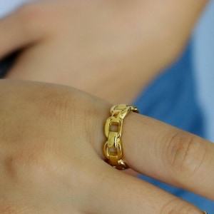 May include: A gold-colored chain link ring. The ring is worn on a finger and has a repeating pattern of rectangular links. The ring is made of a shiny, metallic material.
