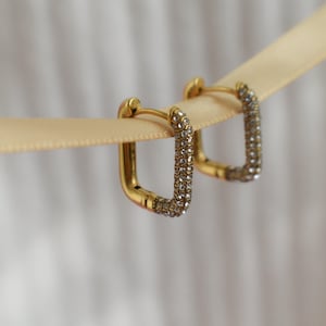 May include: Two gold-tone square hoop earrings, each adorned with tiny, clear crystals. The earrings are presented on a cream ribbon, set against a soft, out-of-focus backdrop.