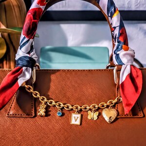 A brown leather handbag with a floral patterned scarf wrapped around the handle, adorned with various charms and a gold chain.