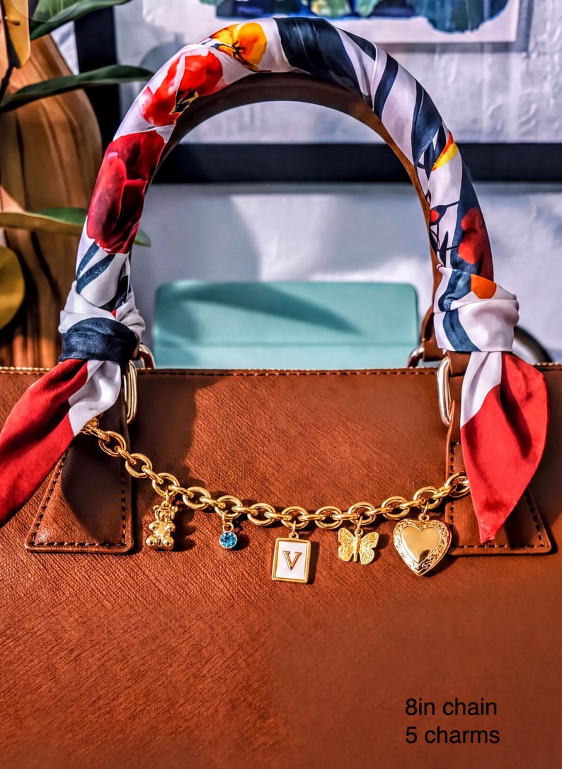 A brown leather handbag with a floral patterned scarf wrapped around the handle, adorned with various charms and a gold chain.