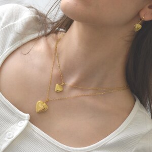 A close-up of a woman wearing a white shirt and two gold heart-shaped pendants on her neck and earrings.