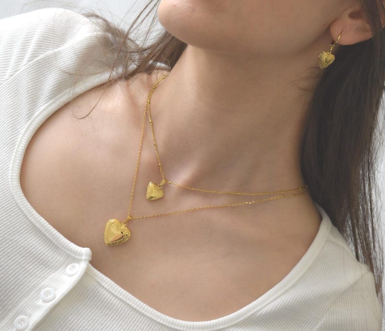 A close-up of a woman wearing a white shirt and two gold heart-shaped pendants on her neck and earrings.
