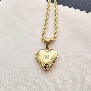 May include: A gold-colored heart-shaped locket necklace with the letter "N" engraved on the front. The pendant hangs from a gold rope chain. The necklace is displayed on a white cloth.