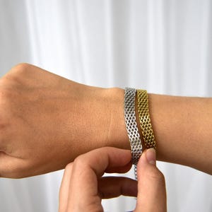 A person's hand is shown adjusting a bracelet on their wrist, with the bracelet being made of metal with a woven pattern.