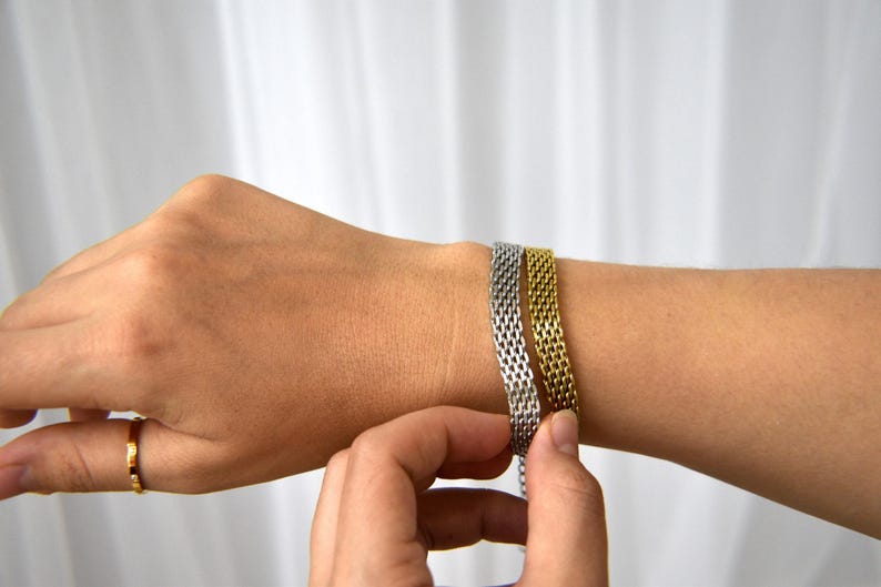 A person's hand is shown adjusting a bracelet on their wrist, with the bracelet being made of metal with a woven pattern.