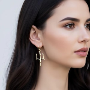 A close-up of a woman's face wearing a pair of gold earrings with a trident design.