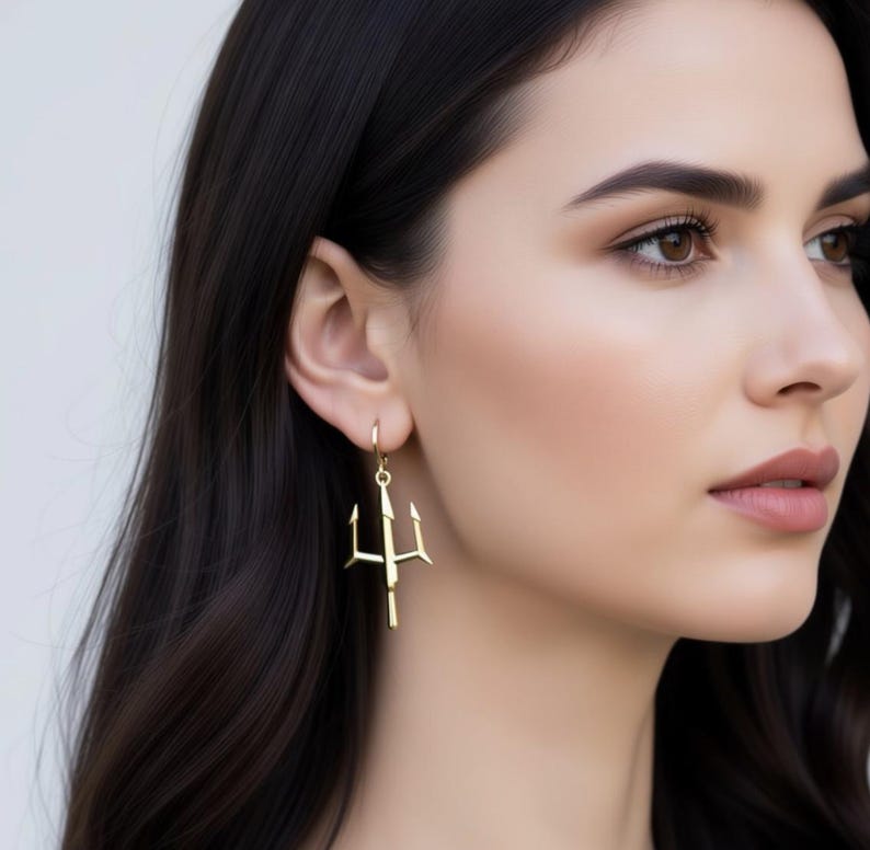 A close-up of a woman's face wearing a pair of gold earrings with a trident design.