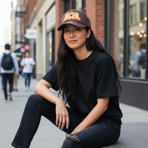 May include: A brown baseball cap featuring an embroidered patch of three light-colored ghosts on the front panel. The cap has a structured crown, a curved brim, and a solid brown fabric construction, providing a simple and functional headwear accessory.