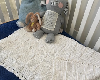 Könnte beinhalten: Eine weiße gestrickte Babydecke mit einem strukturierten Webmuster und einem gewellten Rand. Die Decke liegt auf einer blauen Oberfläche, mit Stofftieren, darunter ein blauer Dinosaurier, ein grauer Elefant und ein braunes Kaninchen.