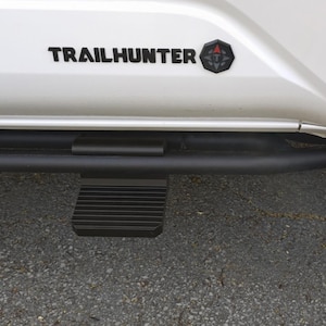 May include: White vehicle side with black "TRAILHUNTER" text and a compass emblem. A black side step with a ribbed step pad is mounted below the door.