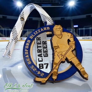 May include: A wooden hockey-themed ornament with a blue and natural wood design. The ornament features a hockey player silhouette, the text "Bradford Blizzard," and the name "Carter Geer" with the number "87." A white and black shoelace is attached.