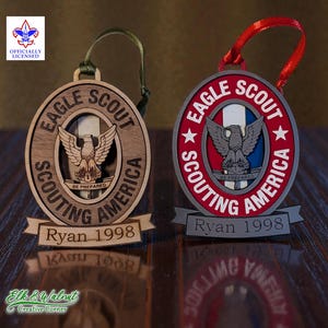 May include: Two Eagle Scout ornaments. One is wooden with a brown finish, the other is metal with red and silver accents. Both feature the Eagle Scout emblem, the text "Eagle Scout Scouting America", and the name "Ryan 1998".