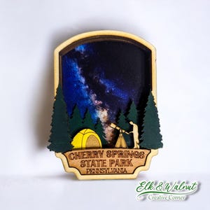 May include: Wooden magnet featuring a night sky scene with a yellow tent, a person looking through a telescope, and trees. The text "Cherry Springs State Park Pennsylvania" is on the bottom of the magnet.
